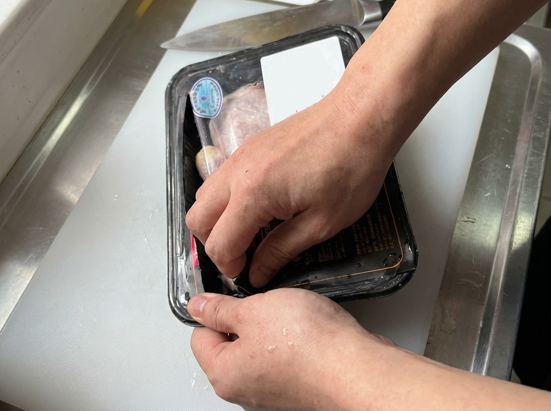白いまな板の上で、包丁の横に置かれたパック入りの生鶏もも肉を開けている手元