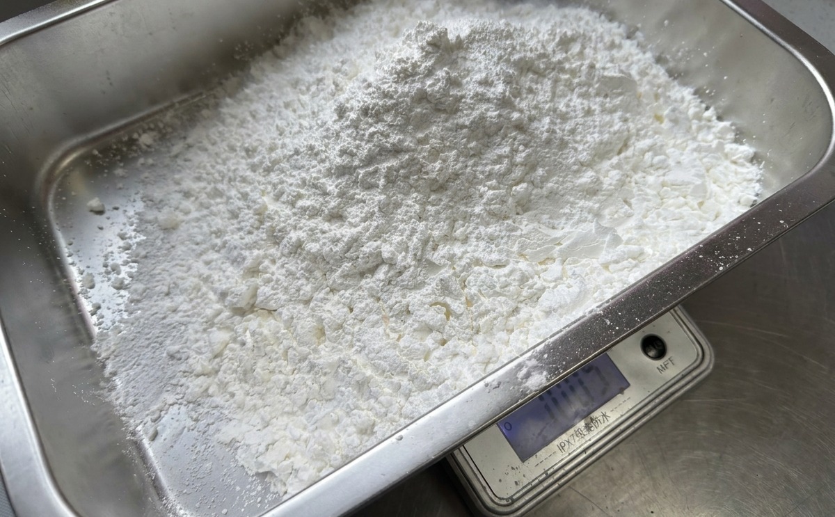 長方形のステンレス製バットの中で、デジタル計りを使って大量の白いコーンスターチを計量している様子