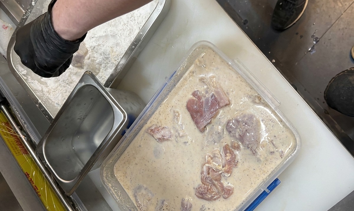 手袋をはめた手で、乳白色のマリネ液に漬かった鶏肉を取り出し、スパイス入りの衣のバットへ移している様子