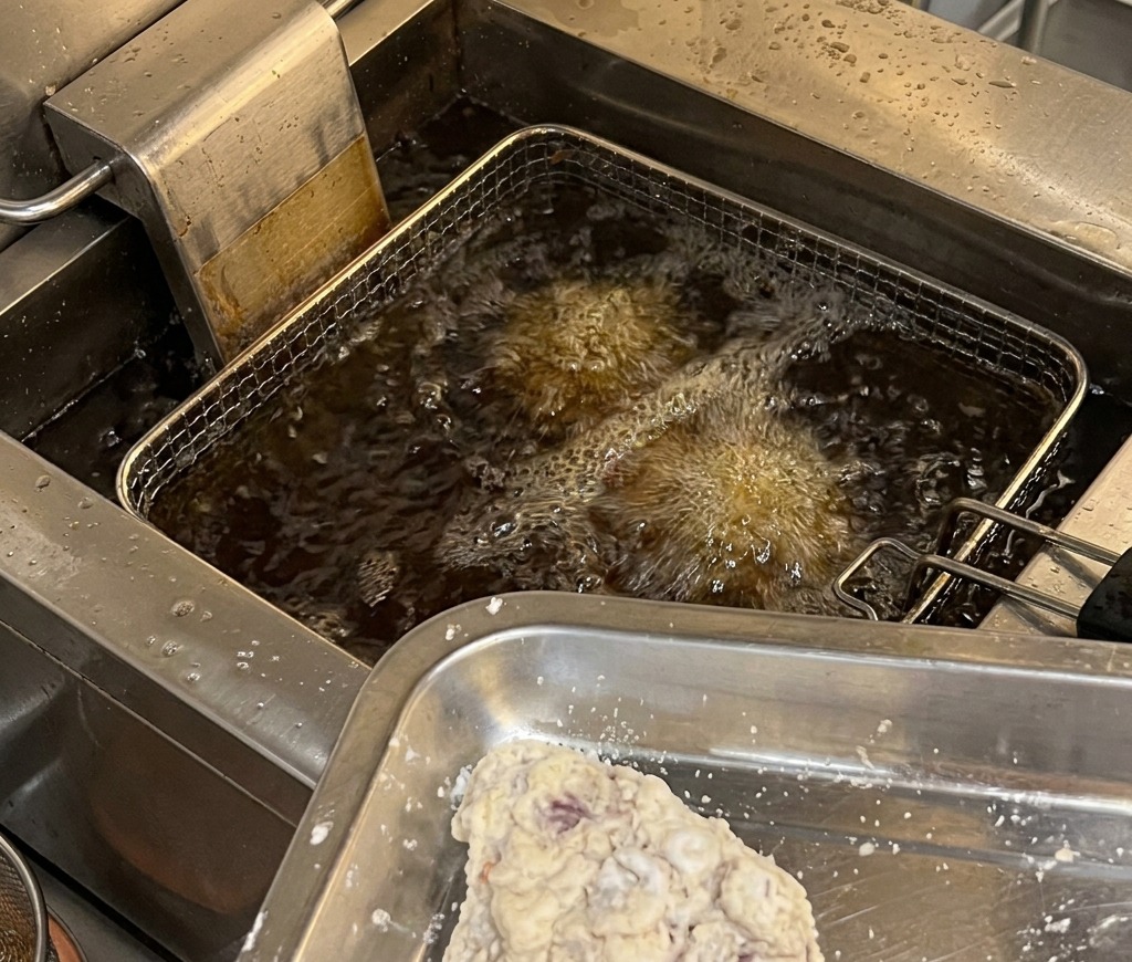 衣をまとった鶏肉が金属製のトレイに並び、高温で泡立つ業務用フライヤーの油に投入されようとしている様子