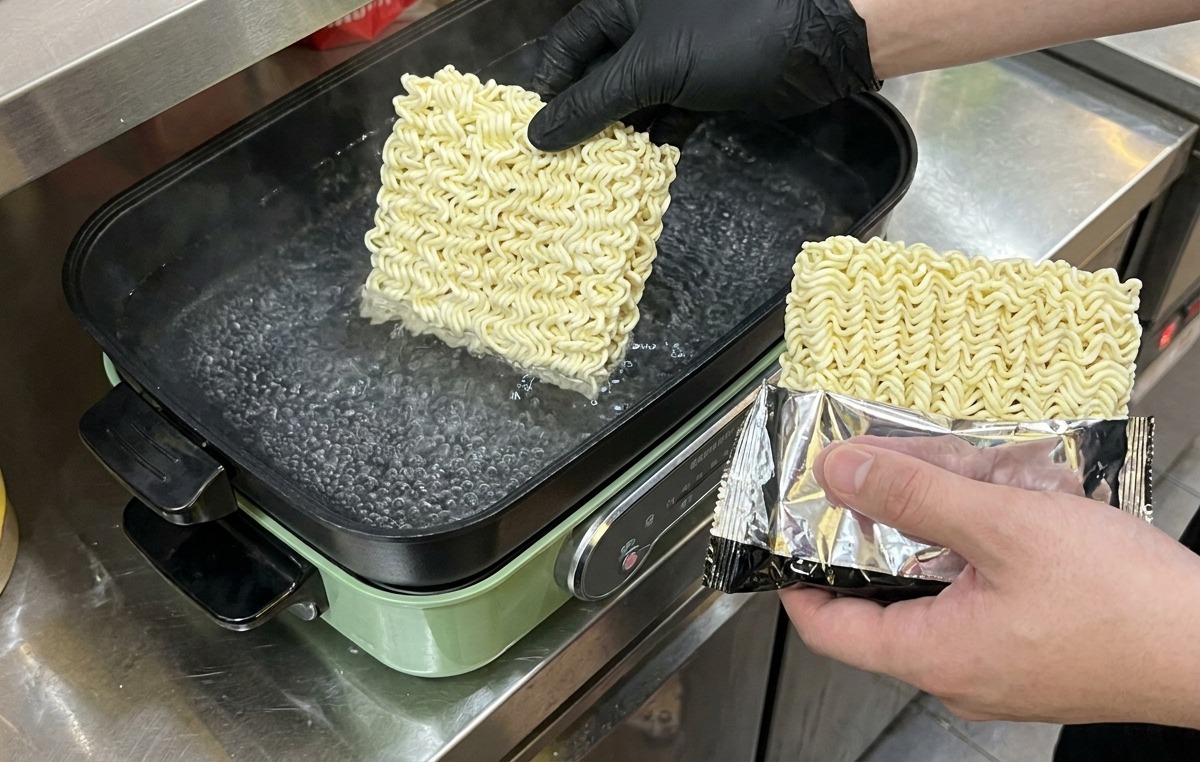 黒い手袋をはめた人が、沸騰した鍋の湯に四角いインスタント乾麺を投入している様子
