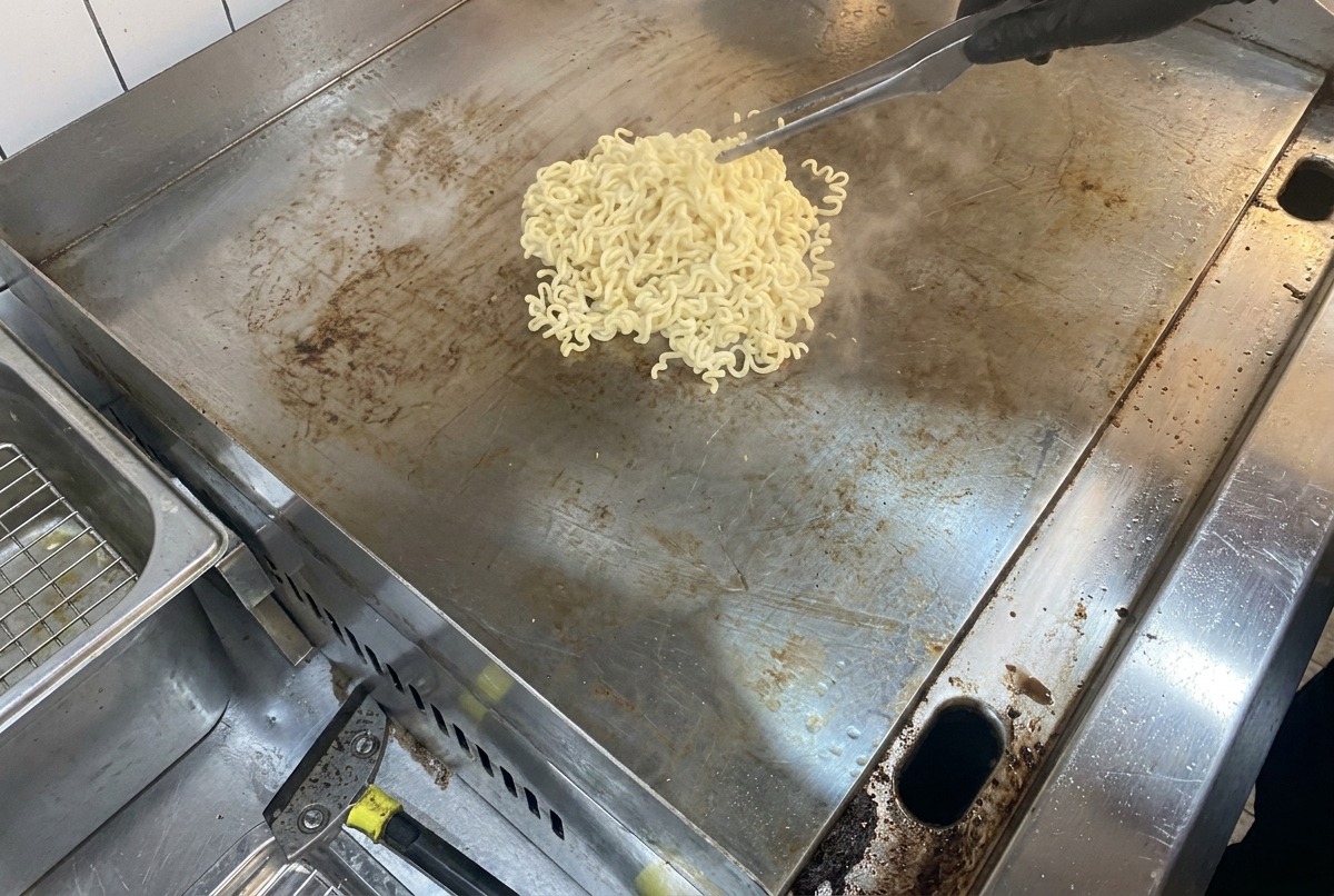 大きな平らな鉄板の上で、湯気を立てる麺と溶け出すバターが絡み合っている様子