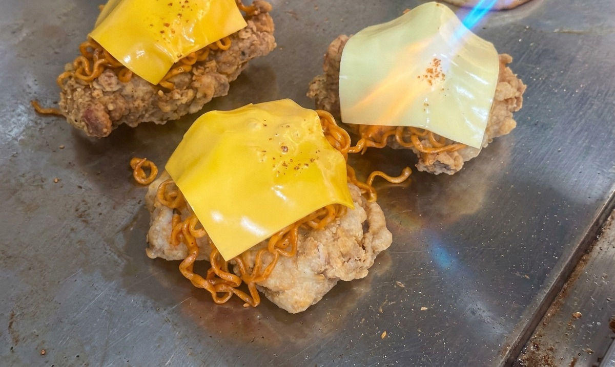 グリルの上で、揚げたてのチキンと激辛麺の層に乗せた四角いチェダーチーズを、バーナーの炎で炙って溶かしている様子
