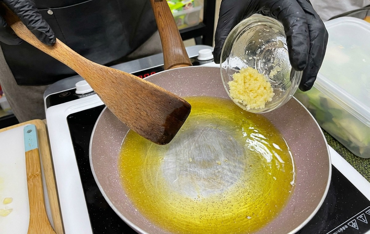黒い手袋をはめた手が、溶けた黄色いバターとオイルの入ったフライパンに、みじん切りにしたにんにくを小さな器から入れている様子