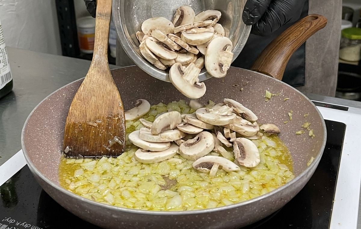 炒めた玉ねぎとにんにくが入ったフライパンに、ボウルいっぱいのスライスされた生のマッシュルームが加えられている様子