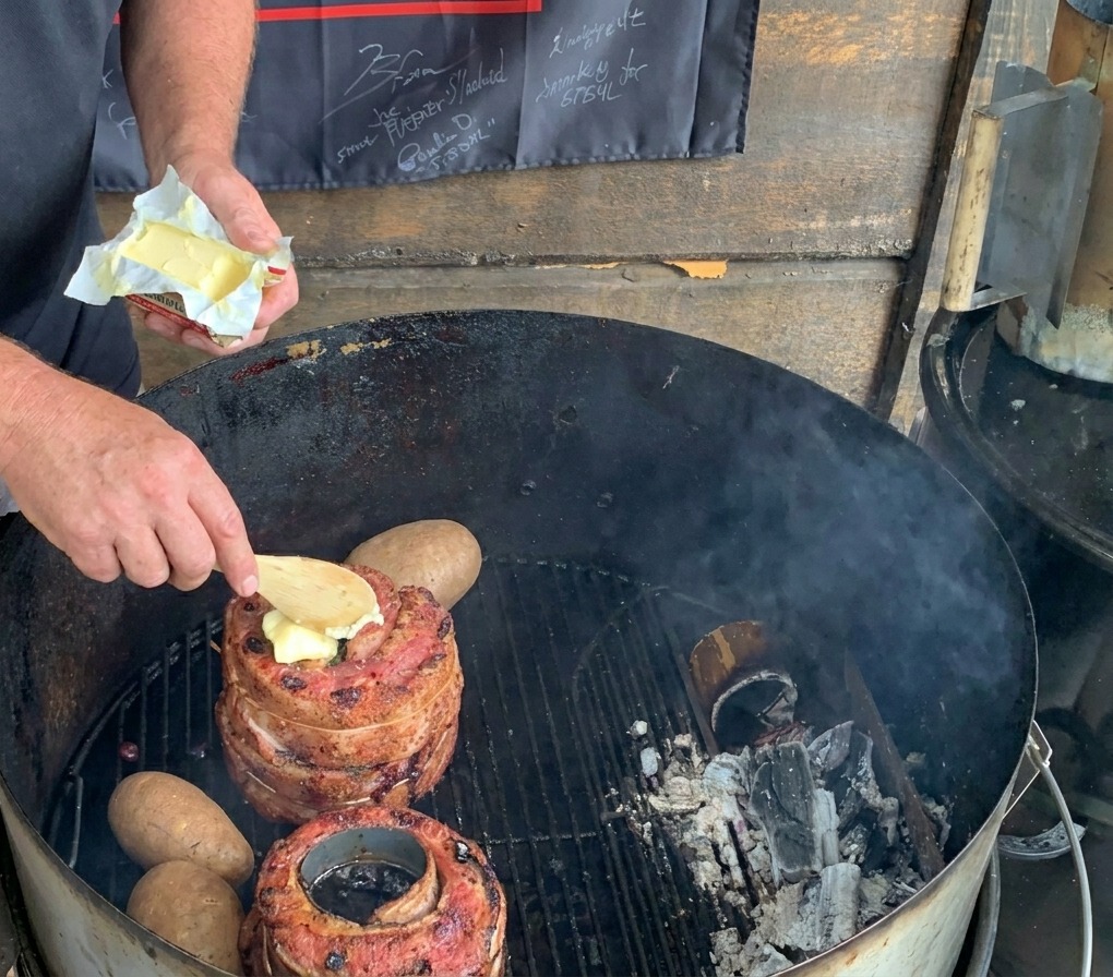 炭火グリルの上で、丸ごとのジャガイモの横にある肉の上のビール缶の中に、木製のスプーンでひとかけのバターを入れている様子。