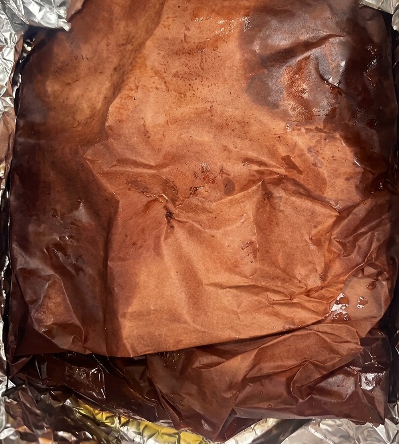 アルミホイルを敷いた容器の中で、肉汁と溶けた脂をたっぷりと吸った濃い色のピーチバッチャーペーパーに包まれた大きなブリスケットの様子。