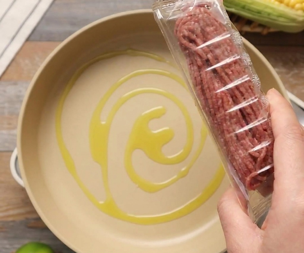 オリーブオイルを引いた明るい色のフライパンの上で、生の牛ひき肉のパッケージを持っている様子。