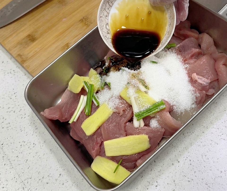 トレイに入った生の豚肉スライス、生姜、長ねぎの上に、小さな器から黒っぽい調味液を注いでいる様子。