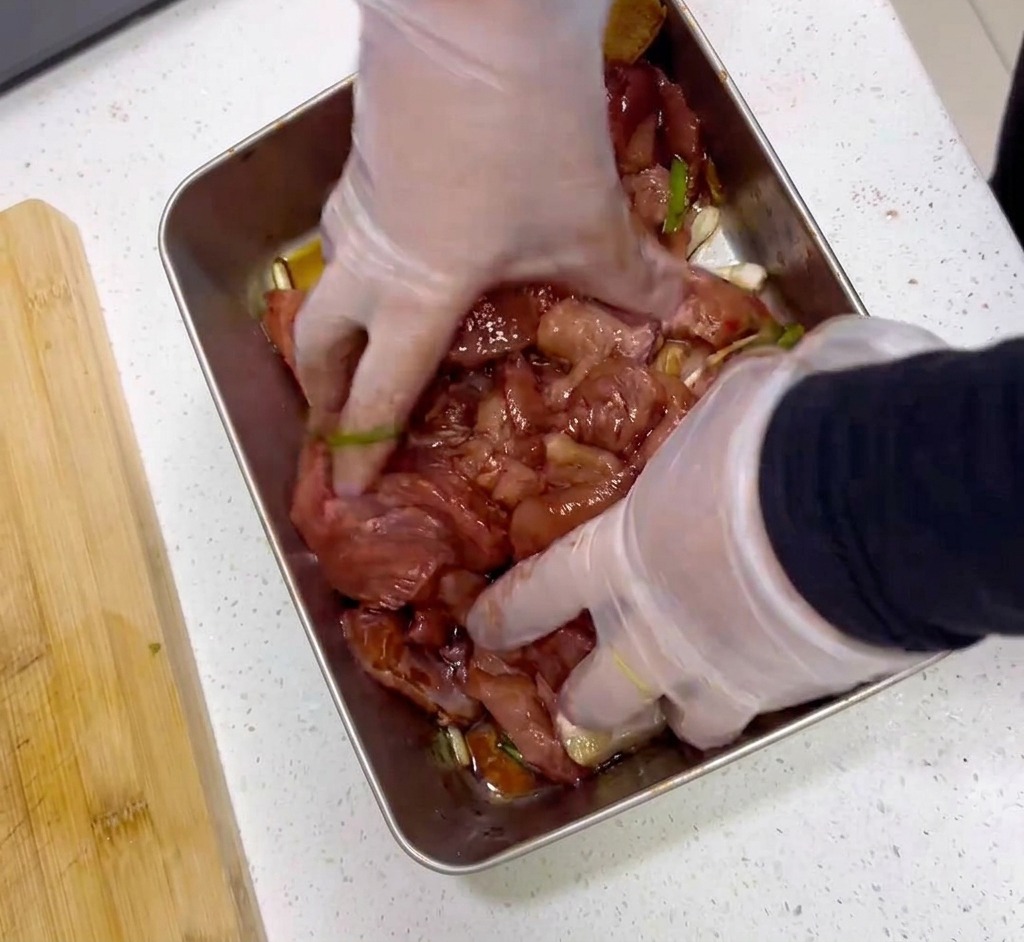 透明なビニール手袋をした両手で、ステンレストレイの中の生肉と濃い赤色の調味液をしっかりと混ぜ合わせている様子。