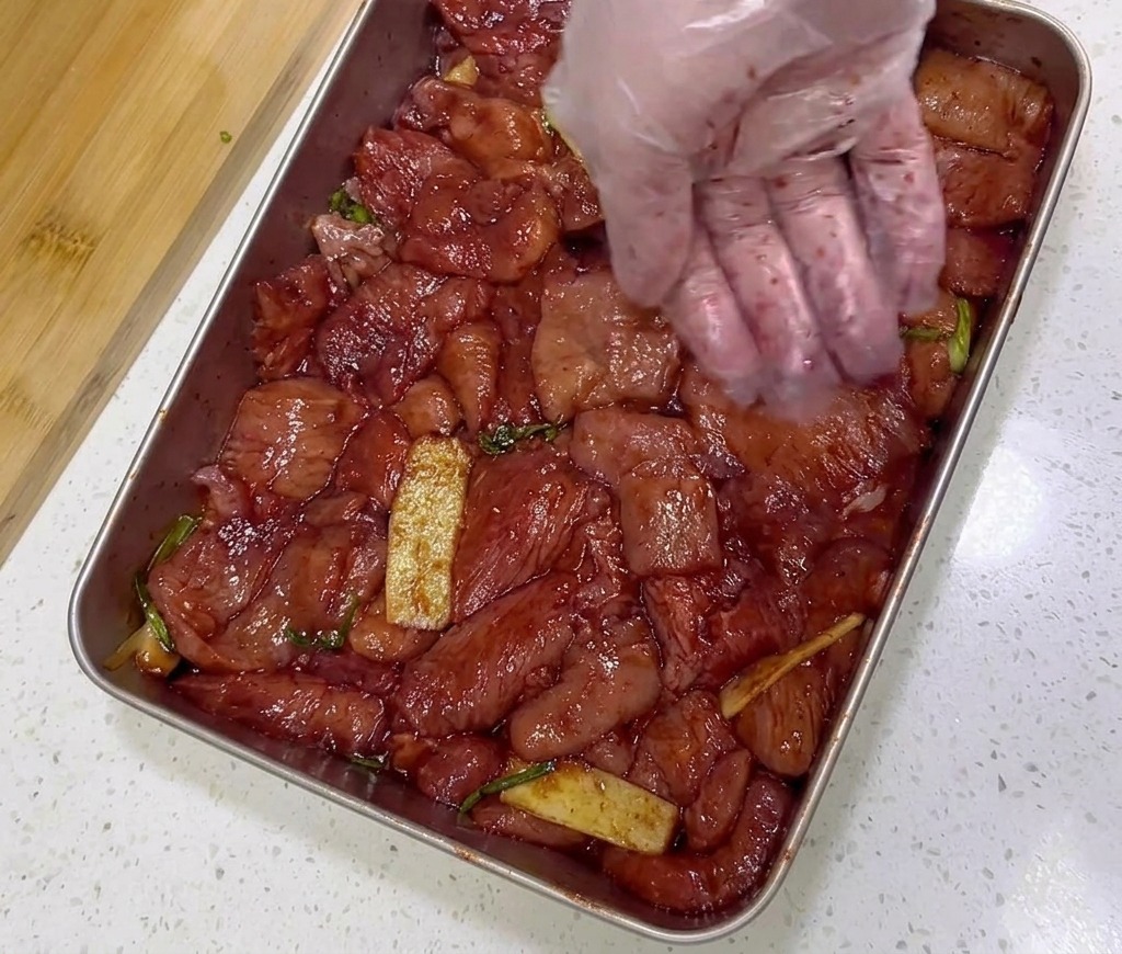 ステンレストレイの中の味付けされた赤い豚肉に、スプーン1杯の粗挽き赤唐辛子を振りかけている様子。