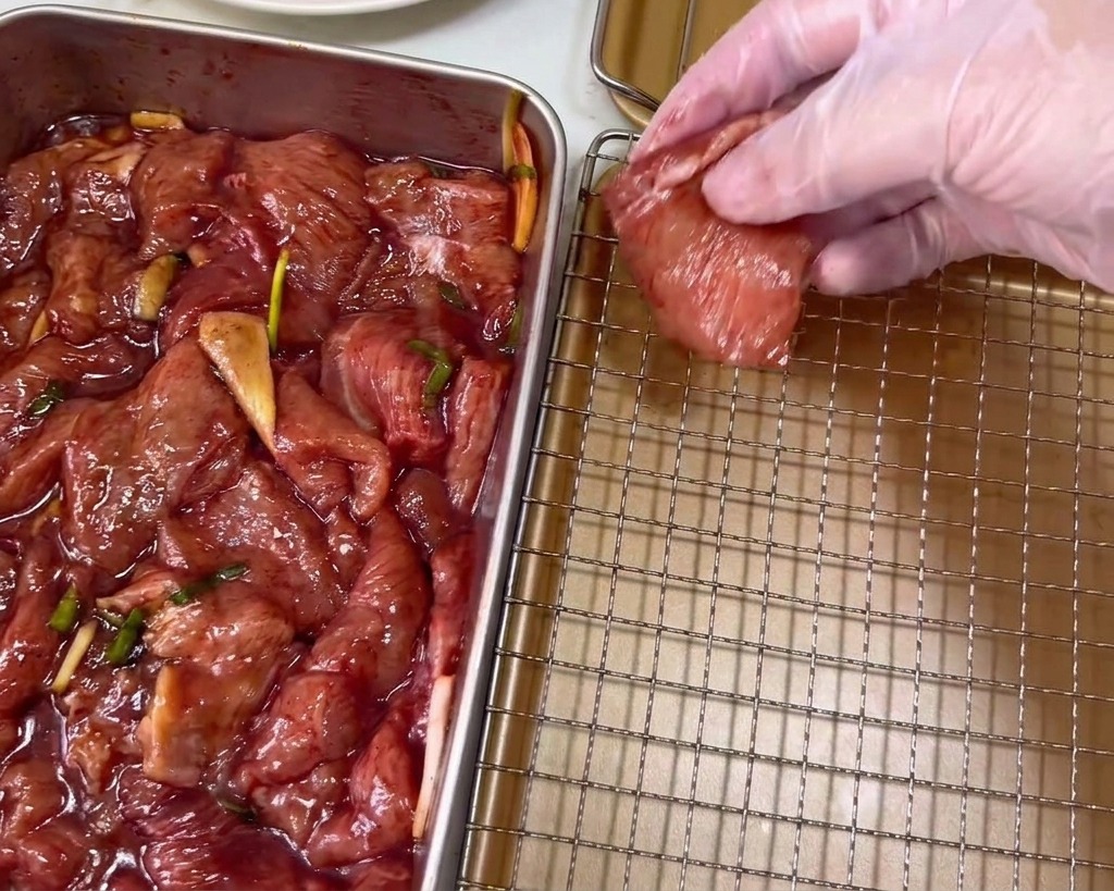 透明なビニール手袋をした手が、濃い赤色に染まった豚肉のスライスを金色のワイヤーラックに並べている様子。