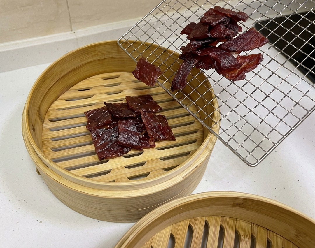 少し硬く乾燥した赤褐色の豚肉を、ワイヤーラックから丸い竹せいろに移している様子。