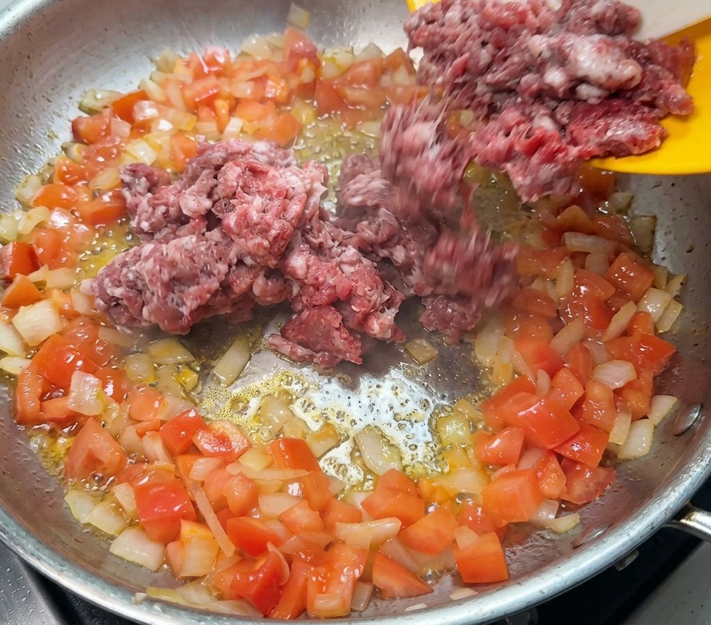 黄色いお皿から、玉ねぎとトマトを炒めたフライパンへ生の牛ひき肉を加えている様子。