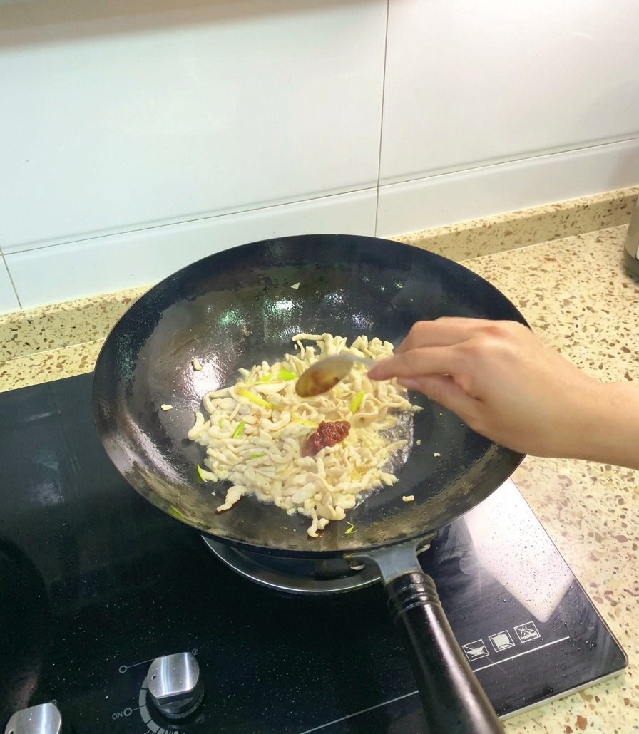 鶏肉と香味野菜が炒められた中華鍋に、スプーンで濃厚な赤色のピーシェン豆板醤を加える様子。