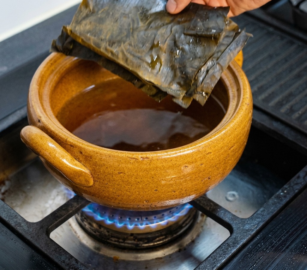 ガスコンロの上の茶色い土鍋に、乾燥した昆布を入れている様子。