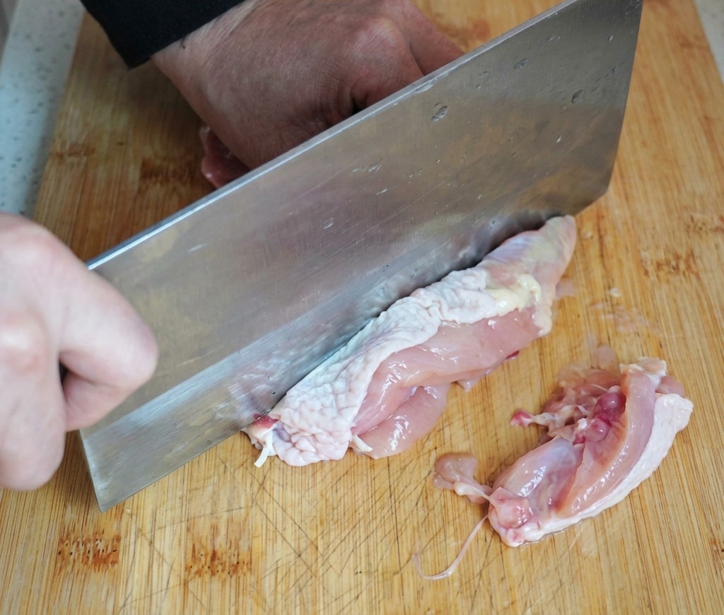木製のまな板の上で、大きめの中華包丁を使って生の鶏もも肉から骨を丁寧に取り除いている様子。