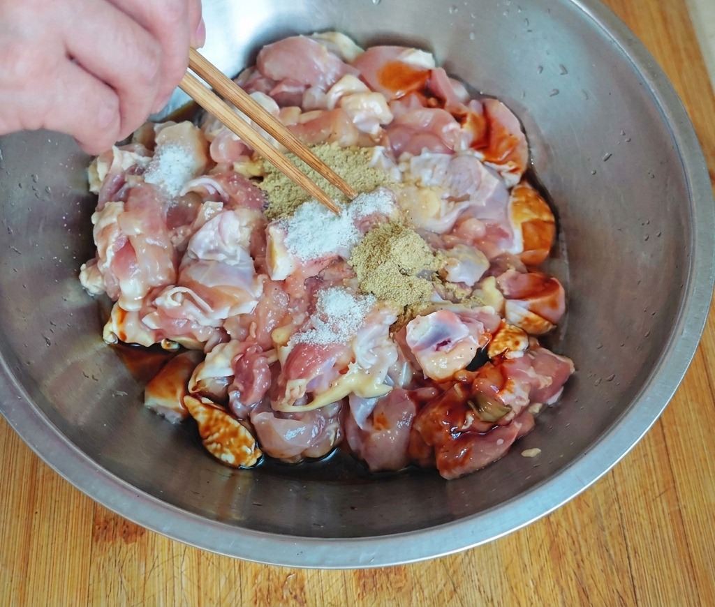 ステンレス製のボウルに入った角切りの生鶏肉に、濃い色の調味料の上から箸を使って白いスパイスを振りかけている手元。