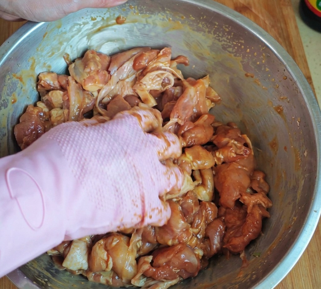 ピンク色のゴム手袋をはめた手が、大きなステンレス製ボウルの中で、下味をつけた鶏肉に澱粉と油をしっかり揉み込んでいる様子。