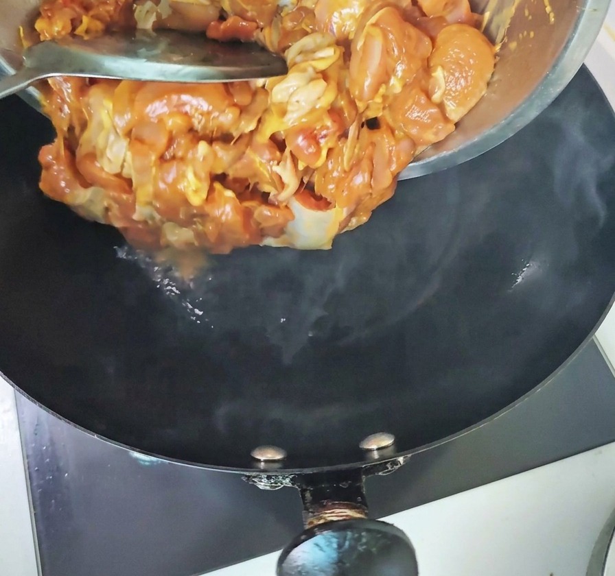煙が立ち上る熱い黒の中華鍋に、金属製のボウルから下味をつけた鶏肉を流し込んでいる様子。