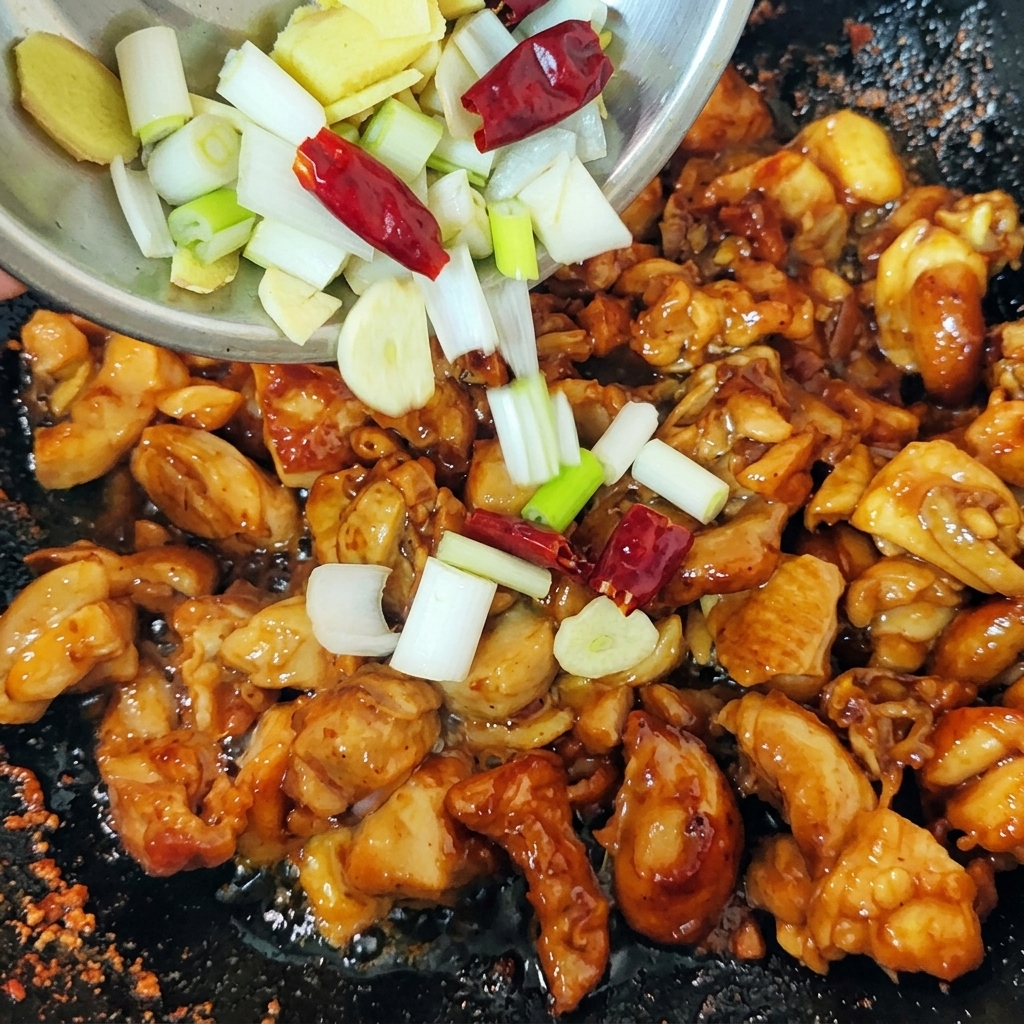 こんがりと焼けた鶏肉が入った中華鍋に、刻んだ長ねぎ、生姜のスライス、乾燥唐辛子が小さなボウルから投入される瞬間。