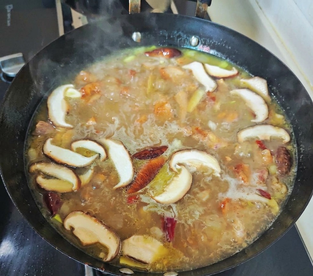 黒い中華鍋の中で、スライスした椎茸と赤唐辛子が、グツグツと泡立つ黄金色の鶏スープと一緒に煮込まれている様子。