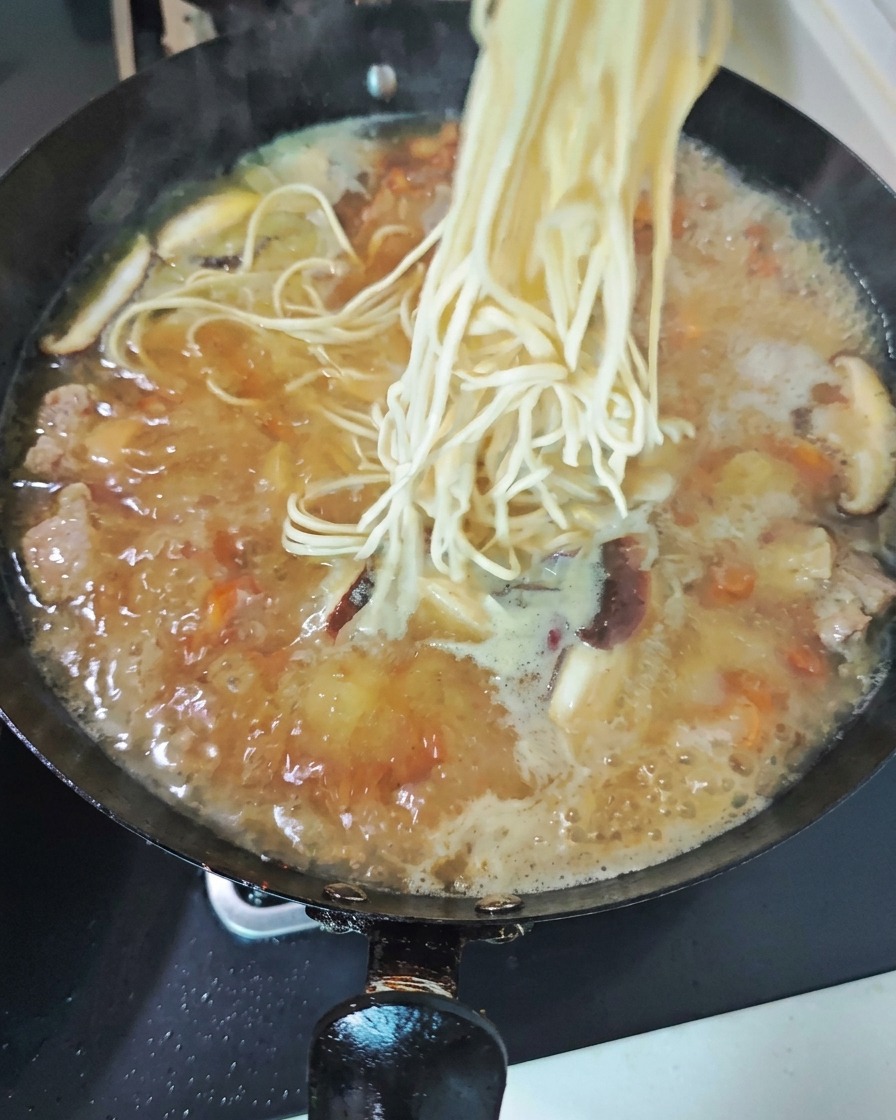 スライスした椎茸がたっぷりと入った、グツグツ煮立つ鶏スープの中華鍋に、ほぐした生の白い中華麺が投入される様子。