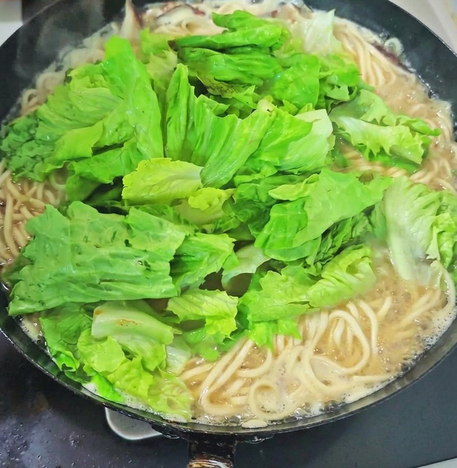 沸騰したスープの中華鍋の中で、茹で上がった麺の上に、たっぷりの新鮮な青菜とにんにくの芽が乗せられている様子。