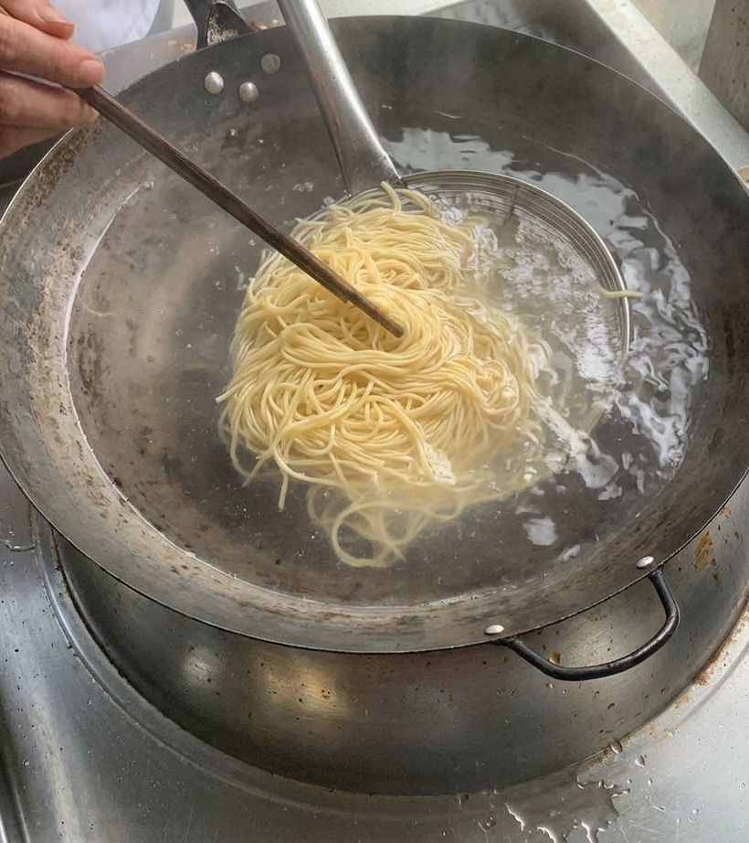 茹で上がった麺を金属製のストレーナーで引き上げ、沸騰した鍋の上で湯切りをしている様子。