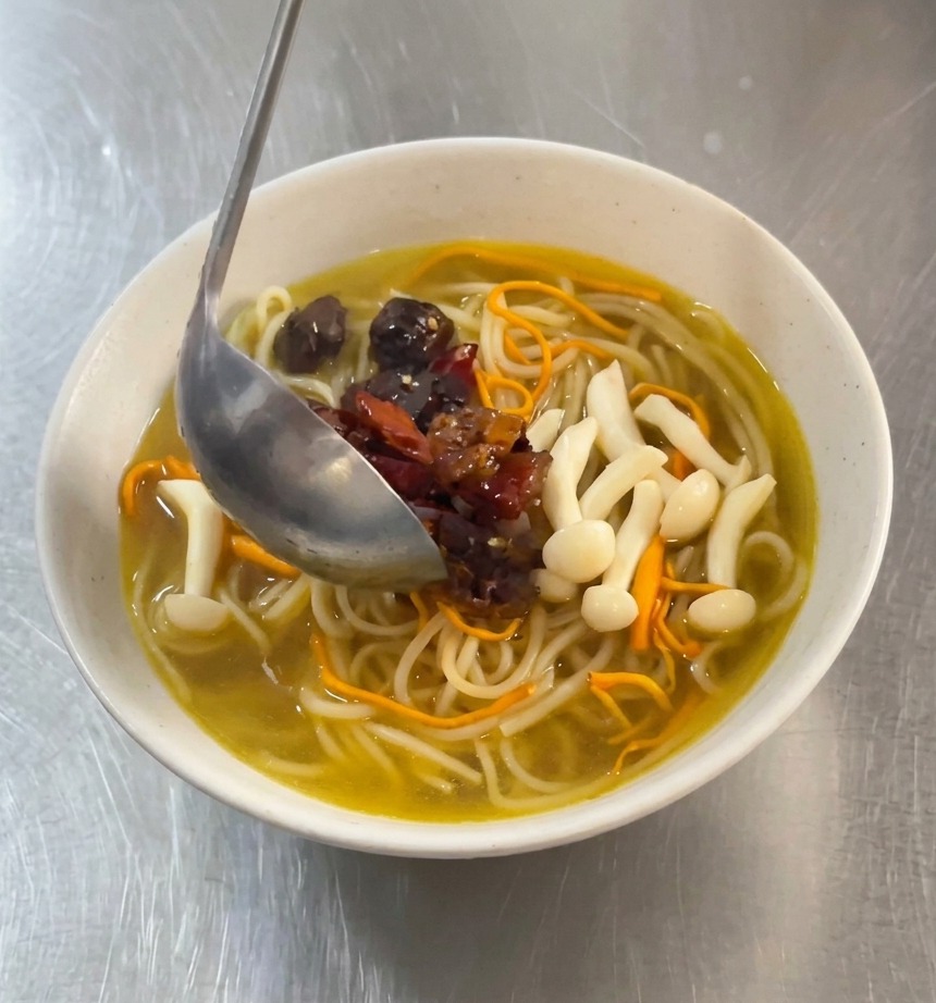レードルを使って、赤く味付けされたピリ辛チキンと乾燥唐辛子を、黄金スープのラーメンの中央に盛り付けている様子。