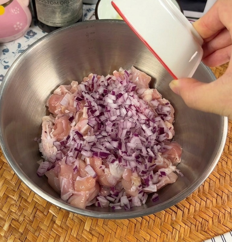 ステンレス製のボウルに入った生の鶏肉のぶつ切りに、みじん切りの赤玉ねぎとニンニクを上から加えている様子。