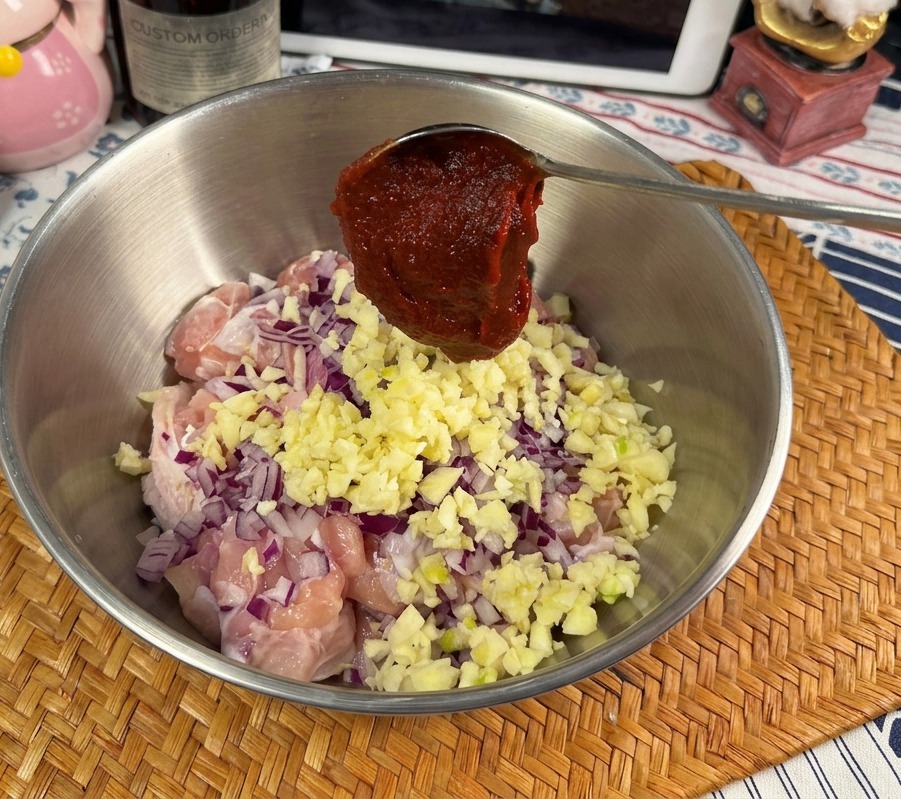 生の鶏肉、にんにく、玉ねぎが入ったボウルの上で、金属製のスプーンに盛られた真っ赤なコチュジャン。