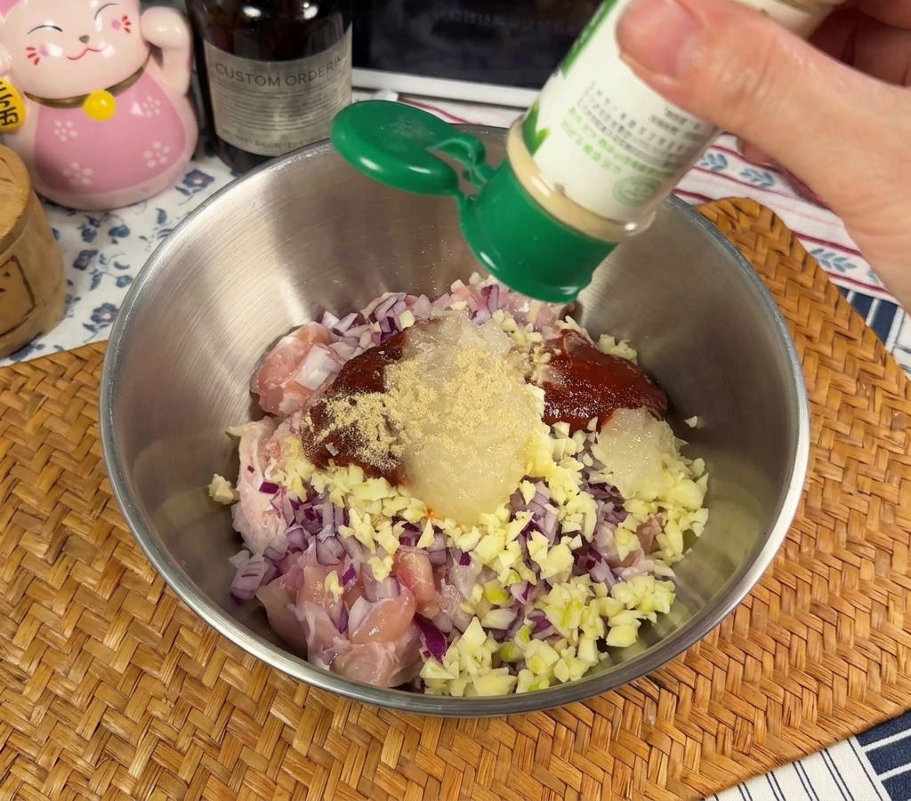 生の鶏肉と刻んだ赤玉ねぎが入った金属製のボウルに、緑色の容器から白コショウの粉末を振り入れている様子。