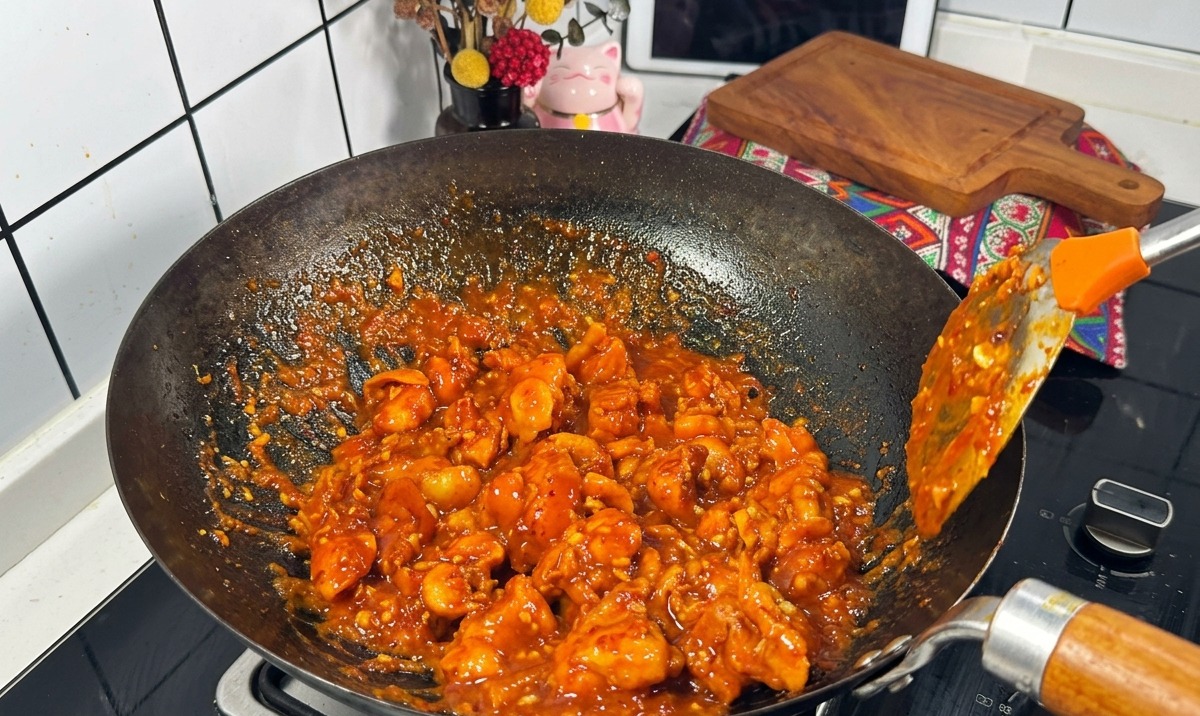 濃厚に沸き立つ赤いソースを纏った鶏肉を、黒い中華鍋の中で金属製のヘラを使って炒めている様子。