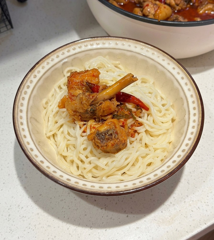 白い麺を入れた陶器の器に、フライパンからピリ辛の鶏肉と赤いスープをたっぷりとかけている様子。