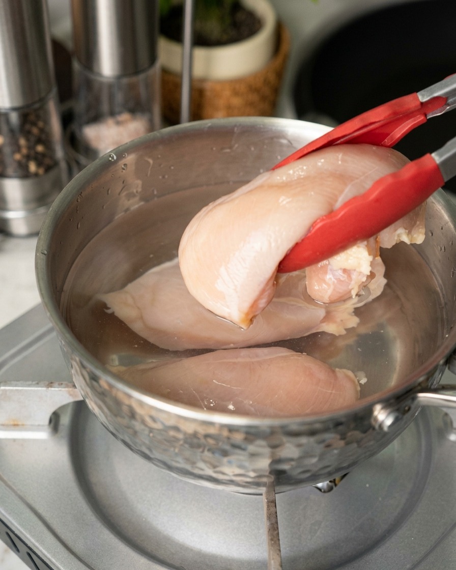 赤い持ち手のトングを使い、生の薄ピンク色の鶏胸肉を水の入ったステンレス製の鍋に入れる様子。