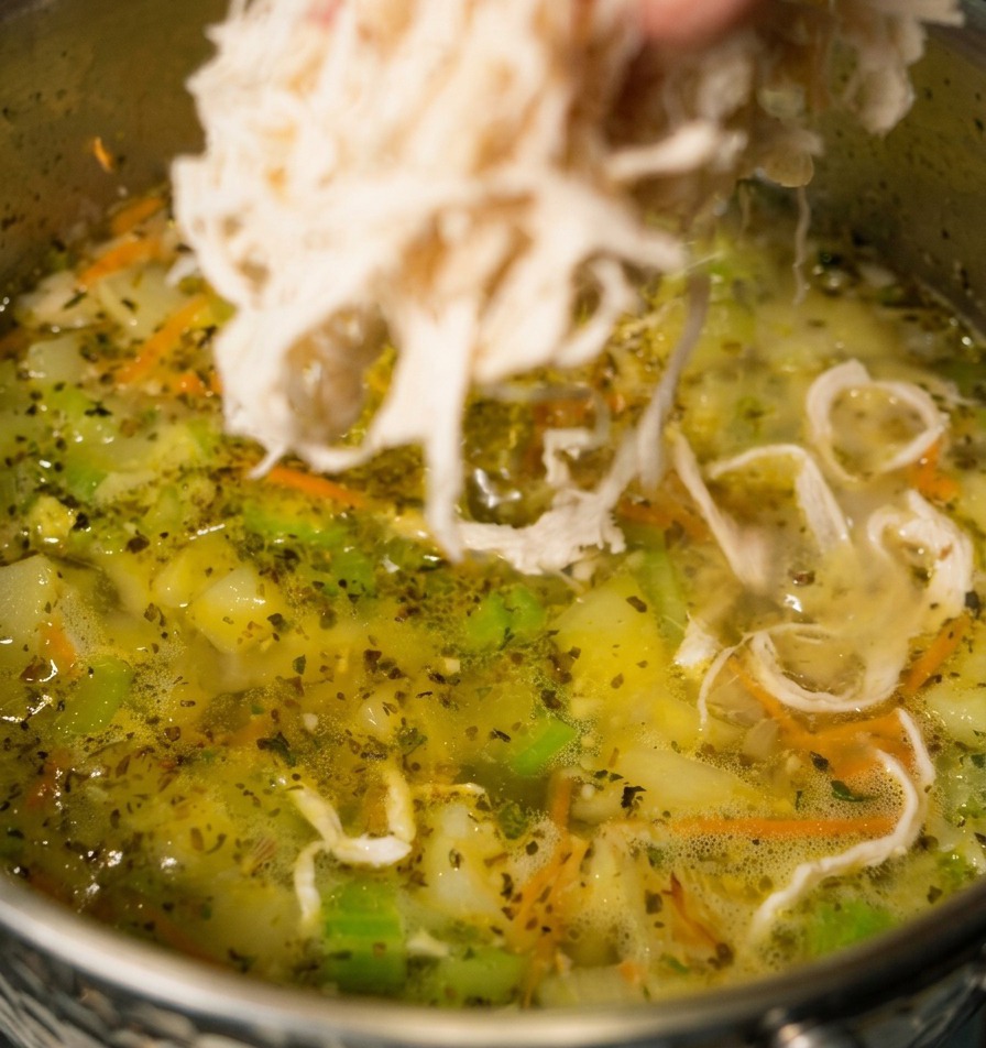 ニンジンやセロリが見える、コトコト煮立つ野菜スープの鍋に、手でほぐした白い鶏肉を入れている様子。