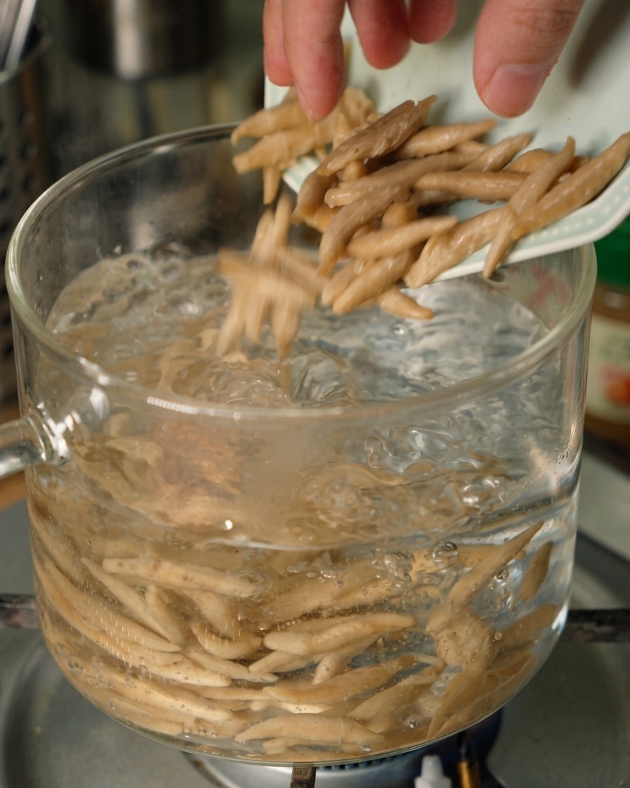 お湯がグラグラと沸騰した透明なガラスの鍋に、端が少し細くなったオートミール麺が小皿から投入されている様子。