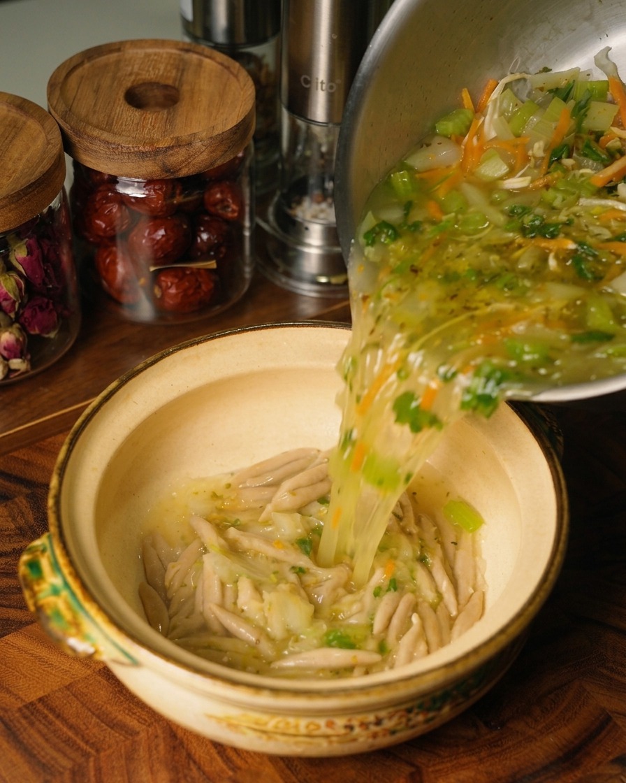 茹で上がったオートミール麺が入った陶器の器に、ステンレス鍋から熱々のチキンと野菜のスープを注いでいる様子。