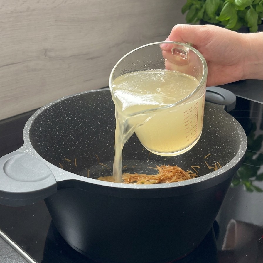 香ばしく炒められた茶色いパスタが入った黒い鍋に、ガラスの計量カップから澄んだチキンスープが注がれている様子。