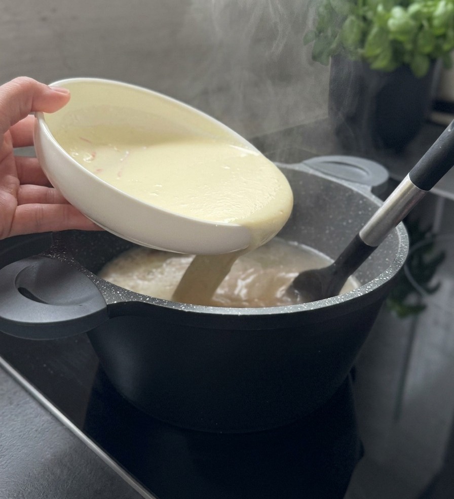 黒いスパチュラで混ぜられている湯気立つスープの鍋に、白いボウルからクリーミーな白いとろみ液が注がれている様子。