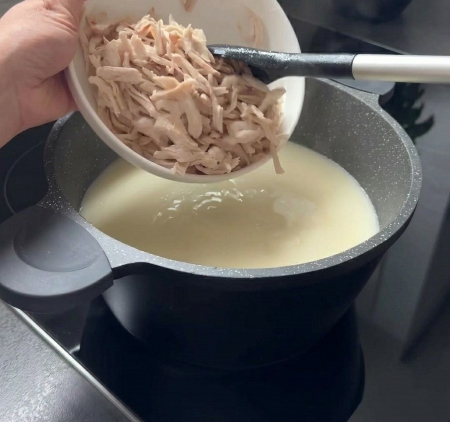 白いボウルからクリーミーな白いスープの鍋に、細かくほぐされた加熱済みの鶏肉が加えられている様子。