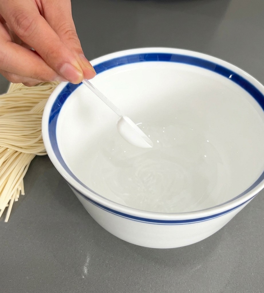 青い縁取りのある白い器に、透明なスプーンで白いスープの素を入れている手元。