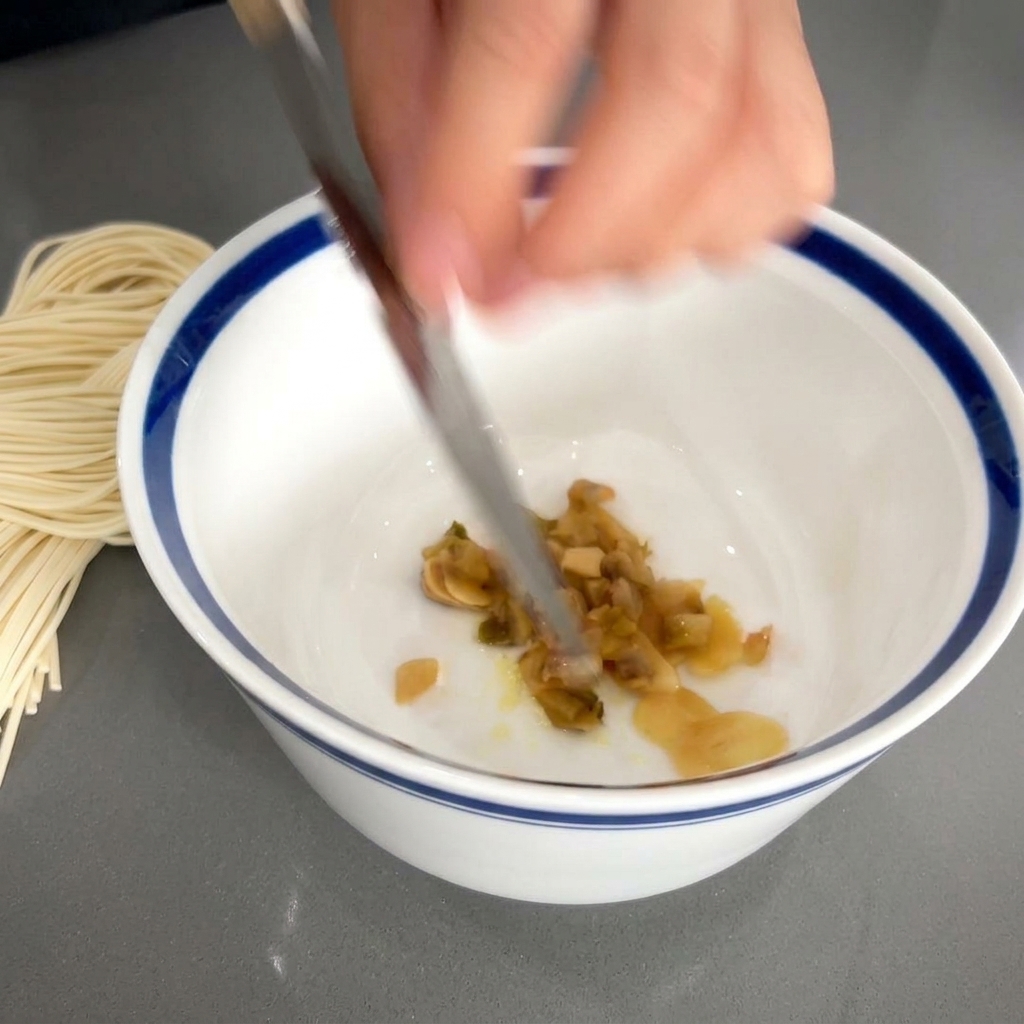 にんにくと調味料が入った白い器に、包丁を使って細かく刻んだ茶色いザーサイを入れている様子。
