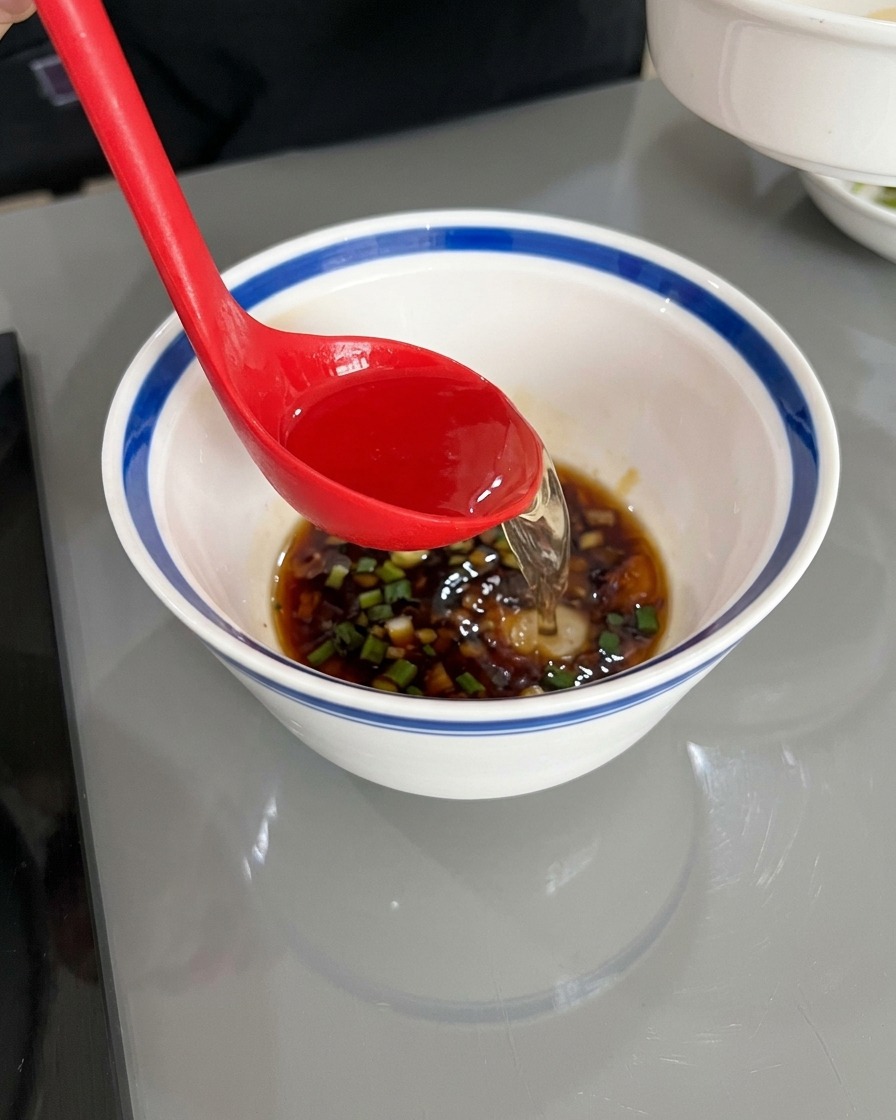 赤いお玉を使い、醤油や油、刻んだ薬味が入った白いどんぶりに、透明で熱いお湯を注いでいる様子。