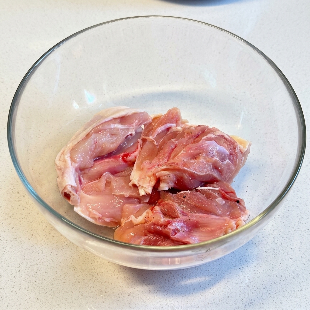 透明なガラスボウルの中で、生の鶏もも肉に塩とこしょうを振っている様子。