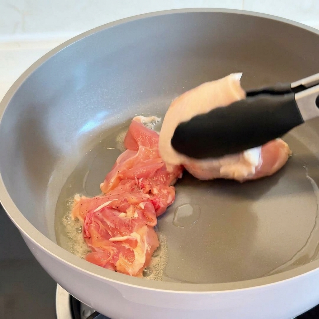 油が温まったグレーのフライパンに、トングで生の鶏もも肉を皮目を下にして並べている様子。