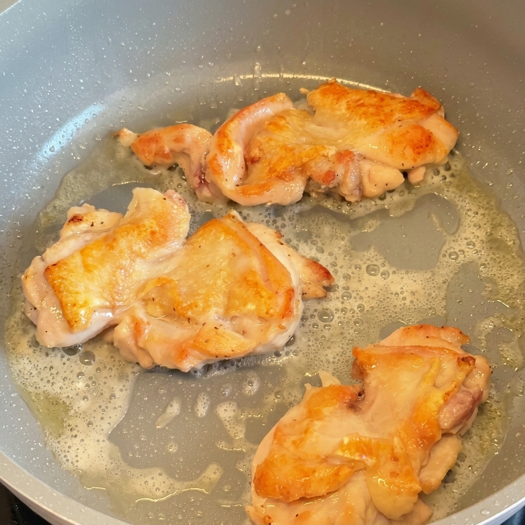 フライパンで3枚の鶏もも肉が焼かれており、ひっくり返されてこんがりと黄金色になった皮目が見えている様子。