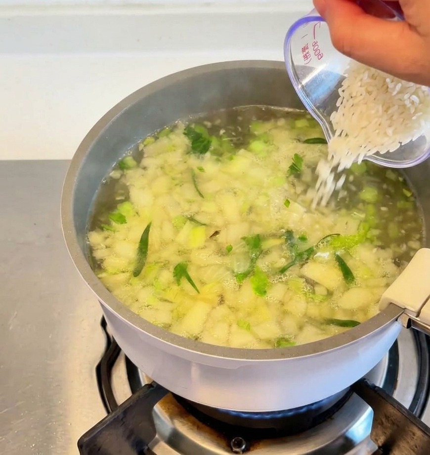 刻んだ野菜やハーブが煮立つスープの鍋に、透明な計量カップから白いお米を注ぎ入れている様子。