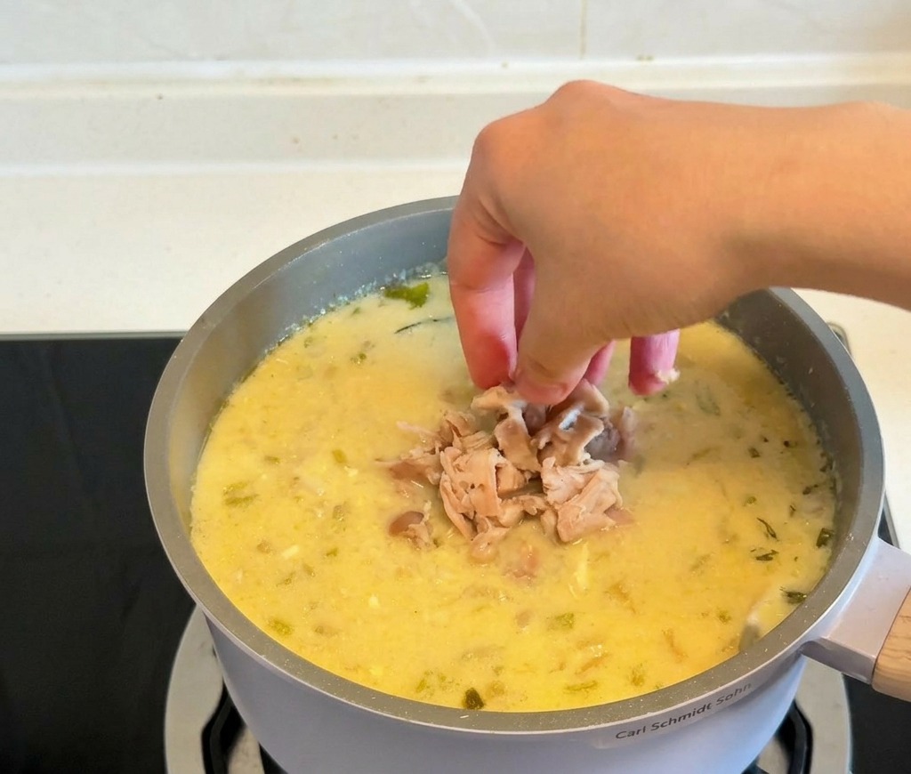 とろみのついたクリーミーな黄色いスープの鍋に、手でほぐした茹で鶏肉を入れている様子。